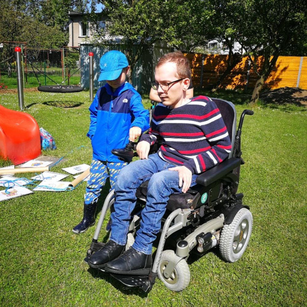 Chlapec si se zájmem prohlíží ovládací joystick elektrického vozíku, na kterém sedí usměvavý lektor na školním hřišti.
